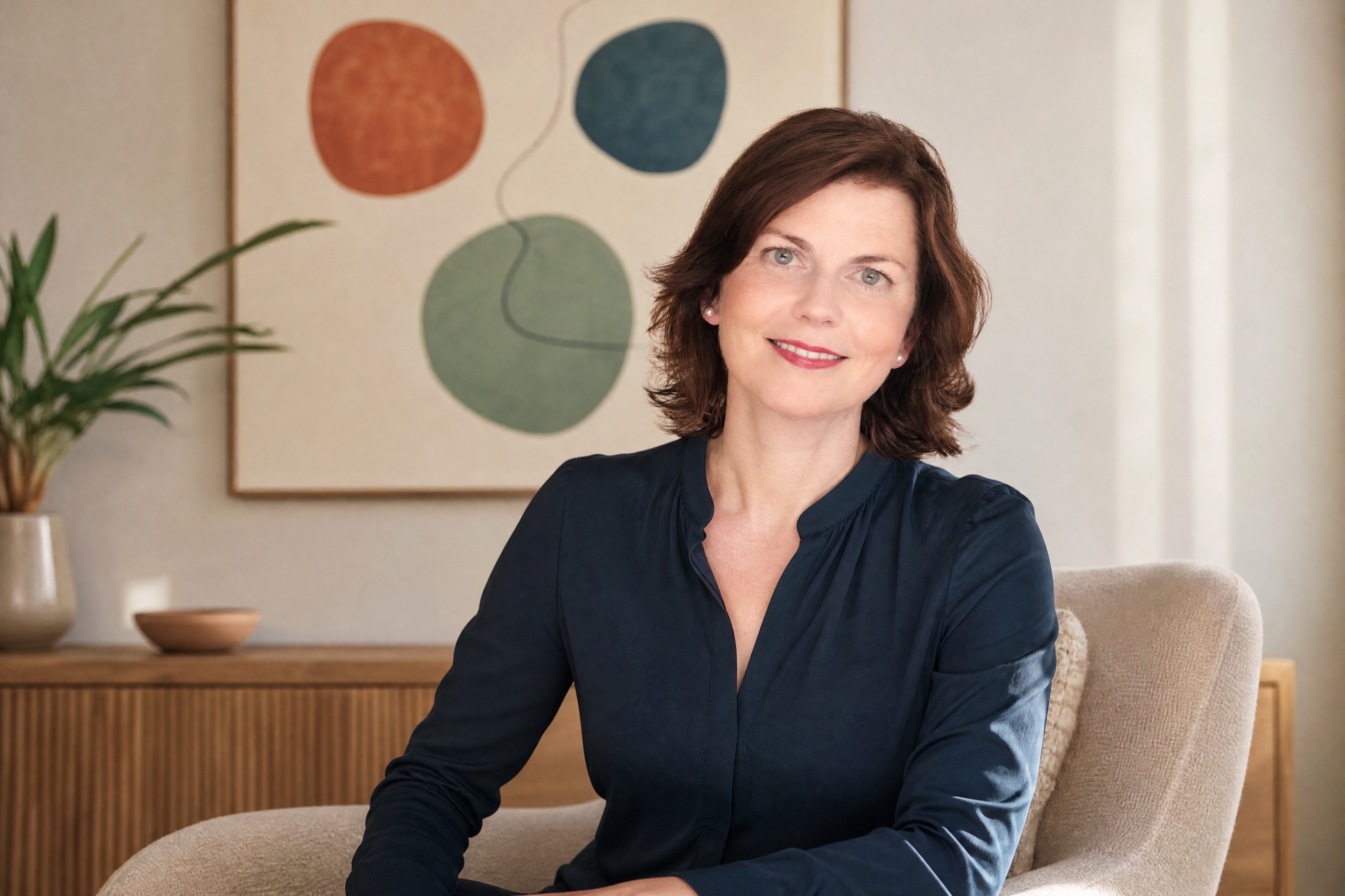 KI-Bild nach echtem Foto: Linda Groß sitzend, mit warmem, offenem Lächeln direkt in die Kamera blickend, im Hintergrund ein Wandbild mit drei Kreisen.
